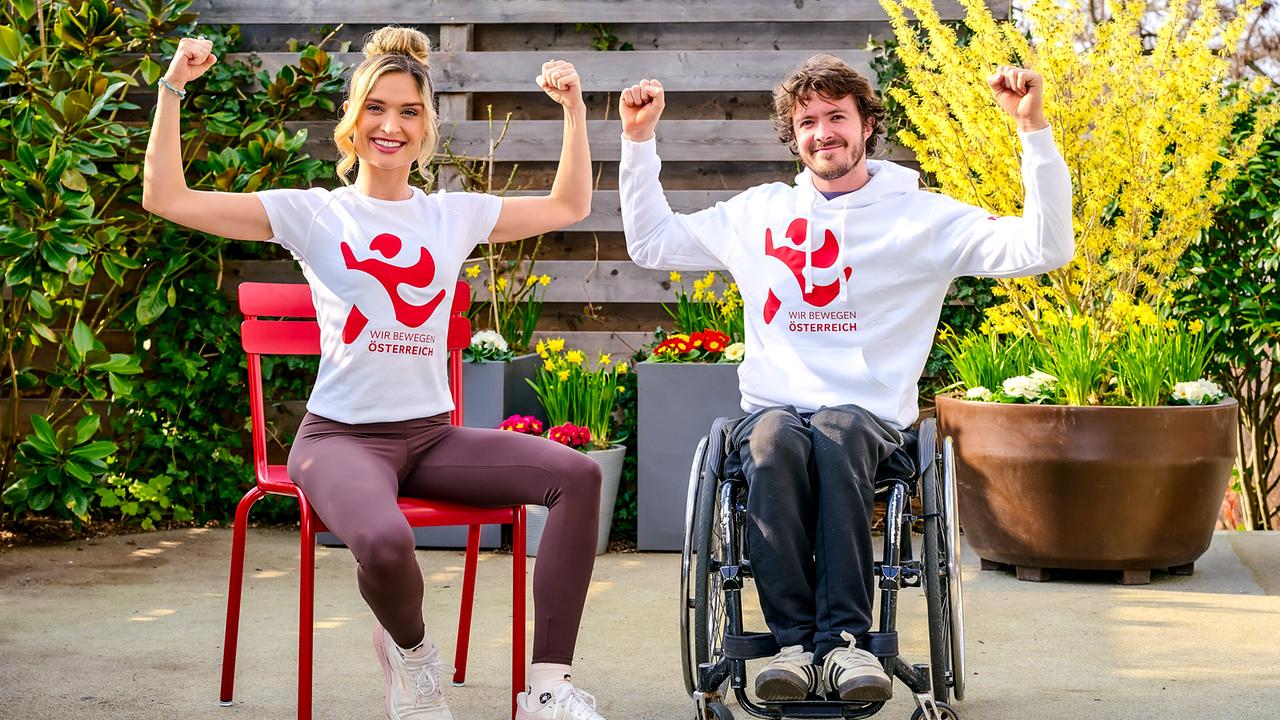 © ORF/Klaus Titzer Corinna Kamper auf einem Stuhl und Nico Langmann in einem Rollstuhl in „WIR BEWEGEN ÖSTERREICH Schule“-Oberteilen sitzen im Freien nebeneinander und heben ihre Arme zum Muskel‑Posieren.
