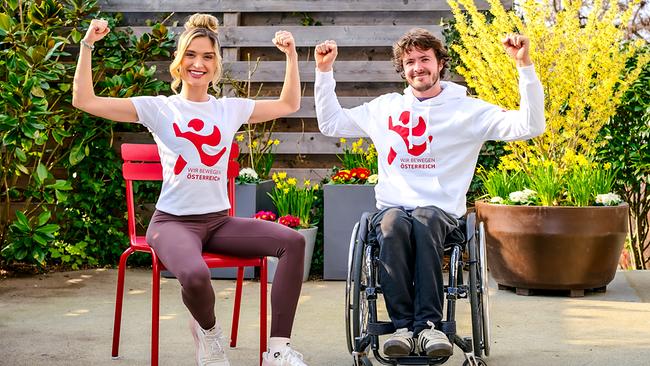 Corinna Kamper auf einem Stuhl und Nico Langmann in einem Rollstuhl in „WIR BEWEGEN ÖSTERREICH  Schule“-Oberteilen sitzen im Freien nebeneinander und heben ihre Arme zum Muskel‑Posieren.