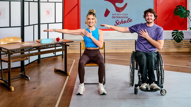 © ORF/Klaus Titzer Corinna Kamper auf einem Sessel und Nico Langmann im Rollstuhl führen gemeinsam eine Oberkörper‑Dehnübung aus, bei der jeweils ein Arm ausgestreckt und die andere Hand auf die Brust gelegt wird - in einem Klassenzimmer-Setting mit der Aufschrift „WIR BEWEGEN ÖSTERREICH Schule“ im Hintergrund.