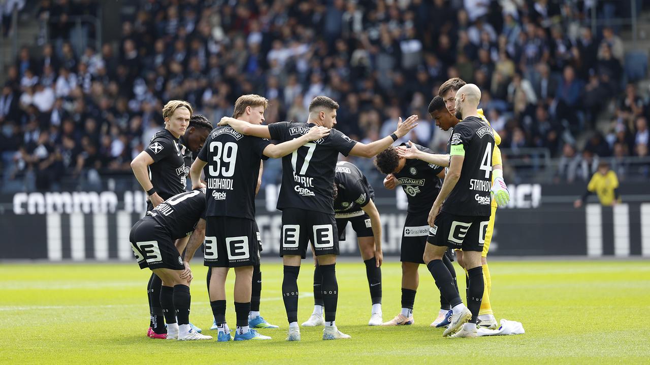 © APA/ERWIN SCHERIAU Die schwarz gekleidete Fußballmannschaft von Sturm Graz bildet einen engen Kreis auf dem Rasen vor Stadionpublikum.