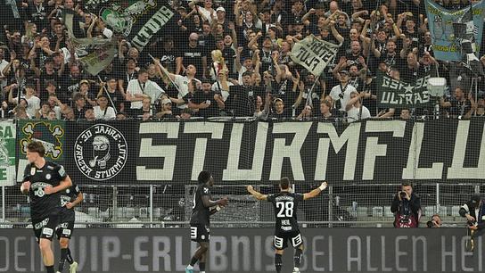 Fußballer Jürgen Heil von Sturm Graz und Kollegen feiern auf dem Feld nach dem 4:0 während dem UEFA-Champions-League Qualifikation, 2. Runde vor Fans, die hinter einer großen Banner mit der Aufschrift „STURM“ jubeln.