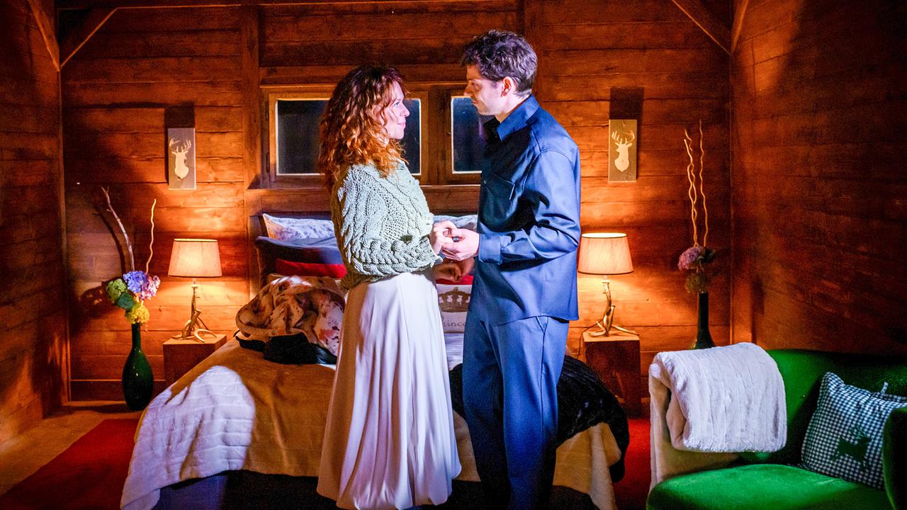 © ORF/ARD/Christof Arnold Johanna Graen und Anthony Paul halten sich die Hände und blicken einander in die Augen in einem rustikalen, romantisch beleuchtenden Zimmer