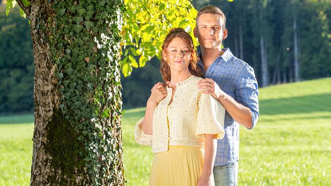 Fanny und Marlon posieren als Paar im warmen Sonnenlicht vor einer grünen Wiese neben einem efeubewachsenen Baum.