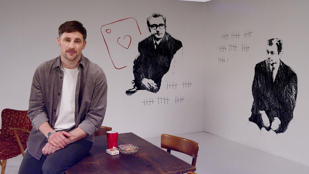 Ferdinand Seebacher sitzt in einem hellen Studio auf einem Holztisch - an der Wand Skizzen von Josef Krista und Josef Angerler