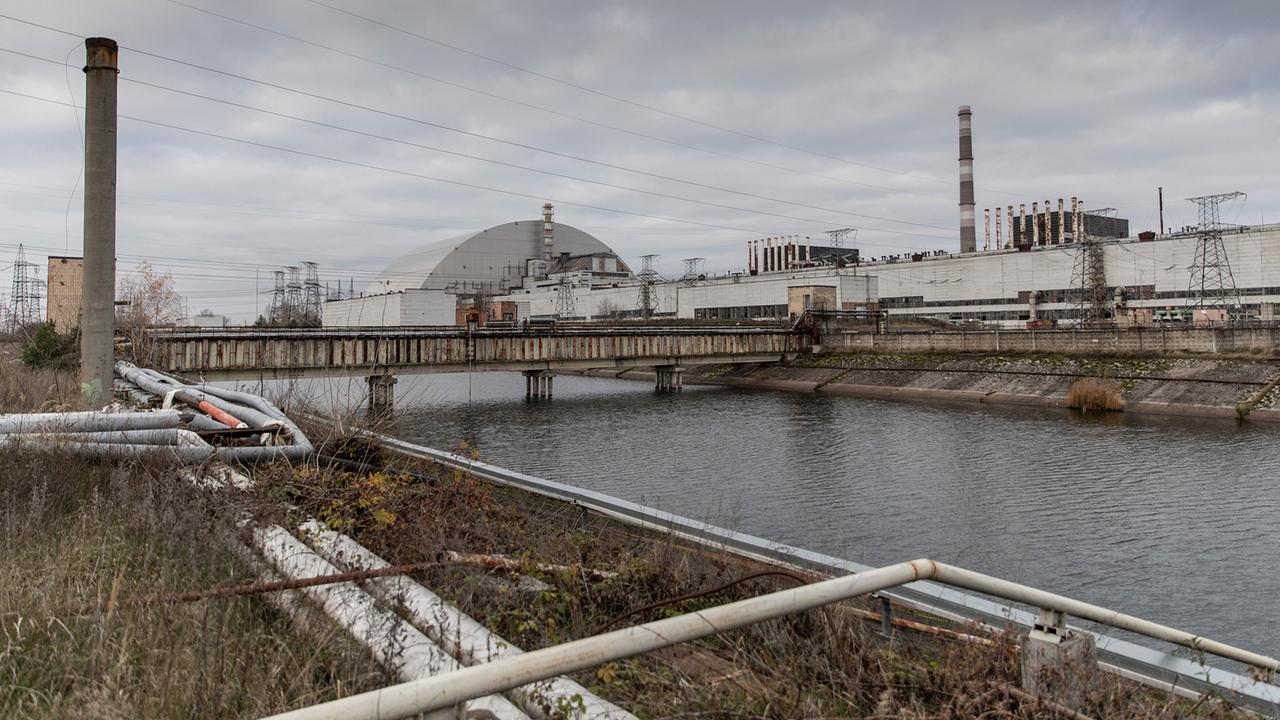 Eine verlassene Industrieanlage mit einem großen Kuppelbau und Schornsteinen in Prypjat an einem Wasserkanal unter grauem Himmel.