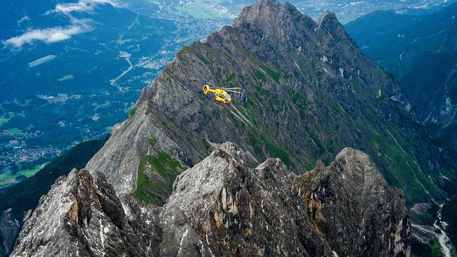 Einsatz der Bergwacht Grainau: Ein gelber Hubschrauber fliegt zwischen zerklüfteten Berggipfeln über einer grünen Tälerlandschaft.