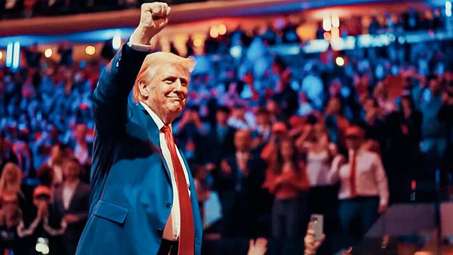 Donald Trump in blauem Anzug mit weißem Hemd und roter Krawatte, er hält seine rechte Faust in die Höhe, das Publikum steht applaudierenden in den Reihen des Veranstaltungssaals