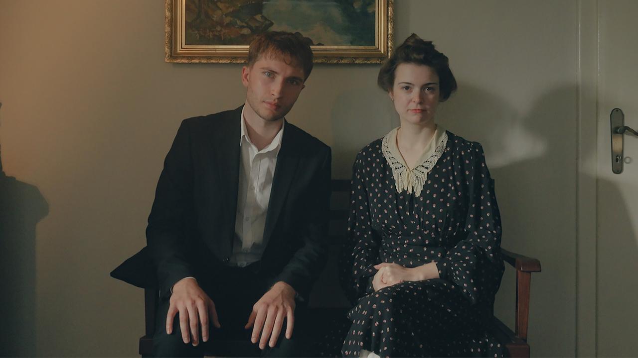 © ORF/HolyScreen Media Reenactment - Emil und Martha Marek sitzen nebeneinander auf einer kleinen Holzbank in einem Biedermeier-Zimmer.