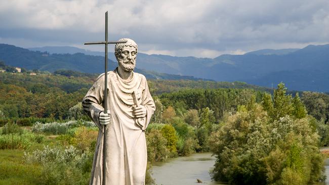 Eine helle steinerne Heiligenstatue mit Kreuzstab vor einer grünen Flusslandschaft und bewaldeten Bergen