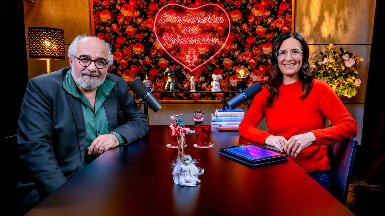 Michael Niavarani und Nina Horowitz sitzen an einem dunklen Holztisch beim ORF-„Liebesg’schichten und Heiratssachen – Der Podcast“