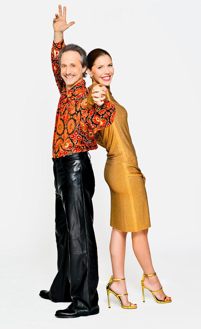© ORF/Thomas Ramstorfer Michael Ostrowski and Victoria Swarovski pose against a white background - Michael in a red floral shirt and black leather pants with an arm raised, Victoria leaning back in a gold dress and yellow high-heeled sandals while they hold hands.