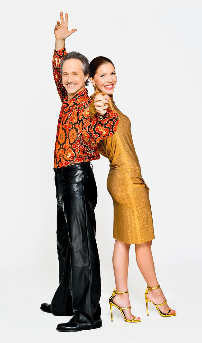 Victoria Swarovski und Michael Ostrowski stehen Rücken an Rücken; Ostrowski trägt ein buntes, retro-blumiges Hemd und schwarze Lederhosen, Swarovski ein goldenes Kleid mit gelben High Heels.