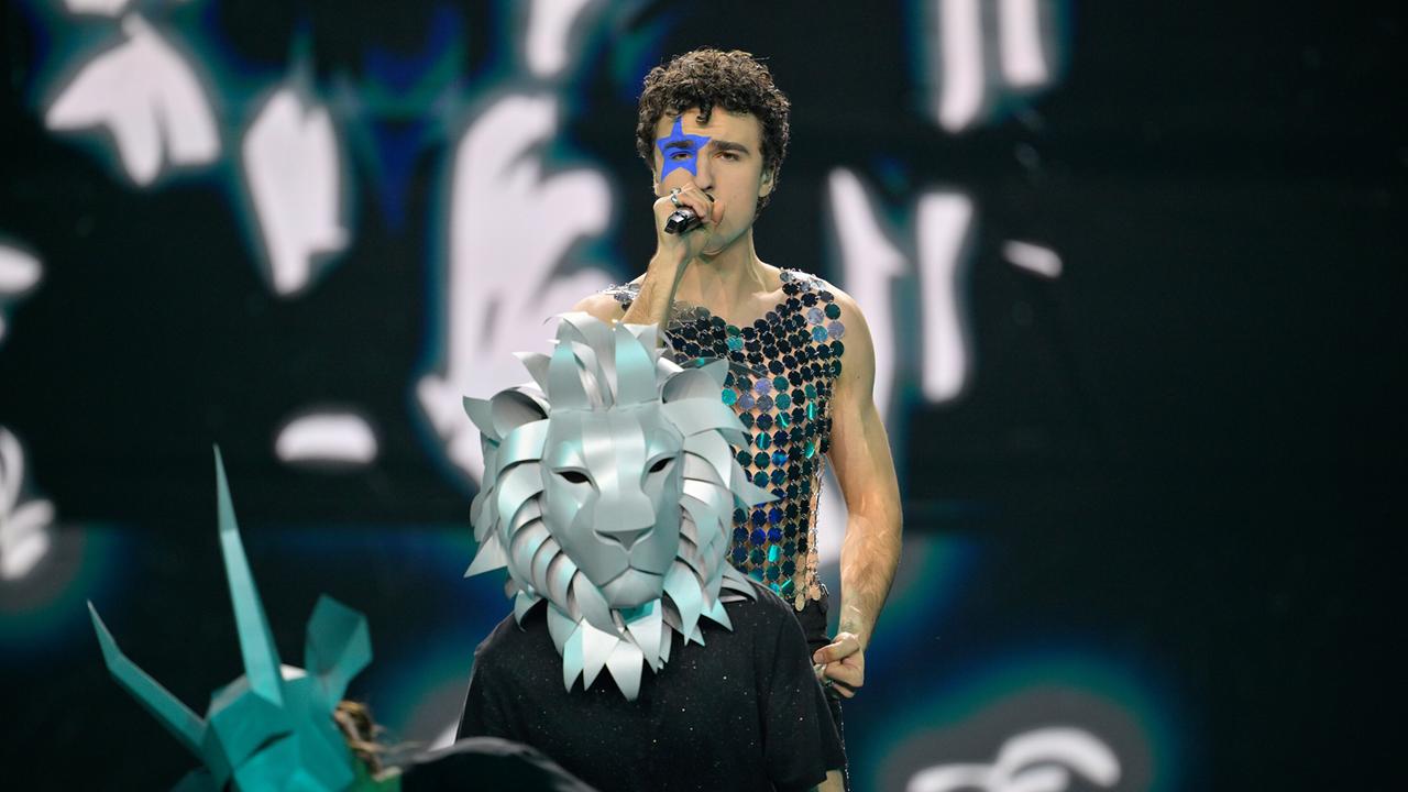 © ORF/Thomas Ramstorfer On a dark stage stands Cosmó in a shimmering costume, in front of him a band member with a silver lion mask and behind that more stylized