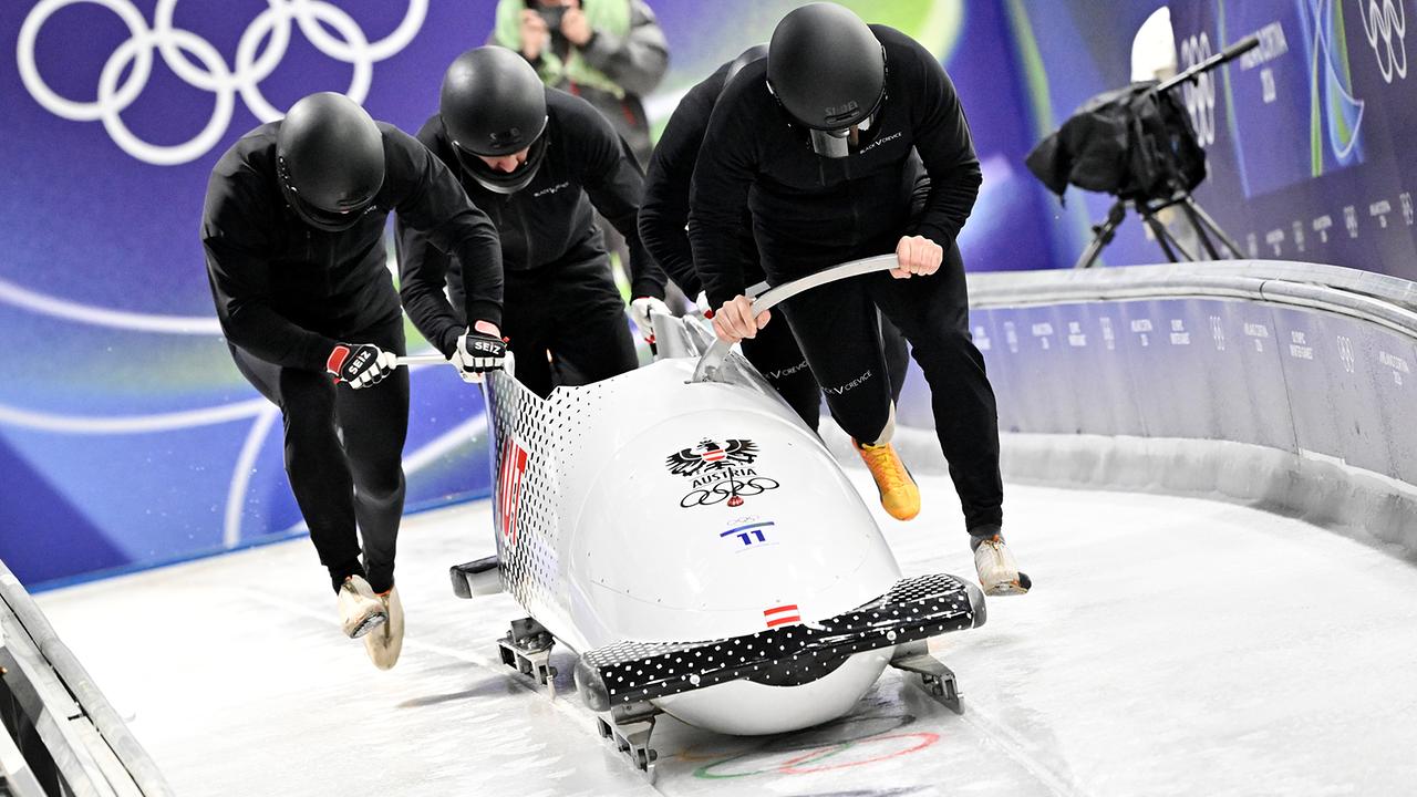 Das österreichische Viererbob-Team mit Markus Treichl, Sascha Stepan, Markus Sammer und Kristian Huber stößt bei den Olympischen Winterspielen seinen Bob am Eiskanal an, im Hintergrund sind die olympischen Ringe zu sehen.