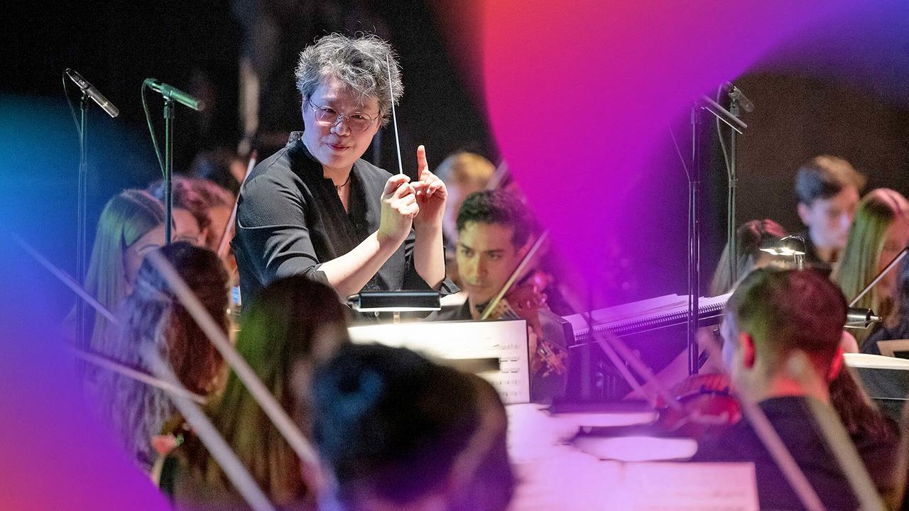 © ORF/ORF Landesstudio Steiermark mit Steirische Kulturveranstaltungen GmbH/Nikola Milatovic Dirigent Mei-Ann Chen leitet das Orchester, umgeben von farbigem Bühnenlicht.