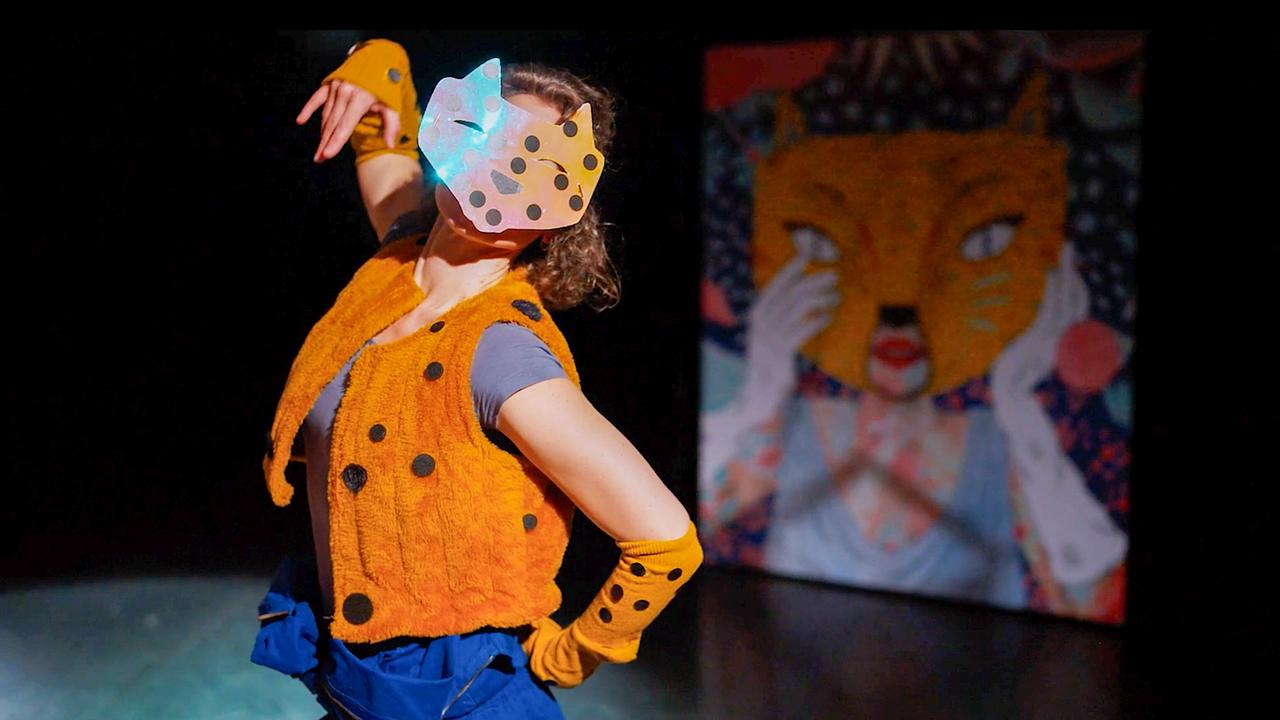 © ORF/Edition 5Haus/Manuel Molzer Eine Tänzerin auf einer Bühne, die eine gepunktete Tiermaske und ein orangefarbenes Kostüm trägt, bewegt sich in einer ausdrucksstarken Pose auf einer dunklen Bühne.