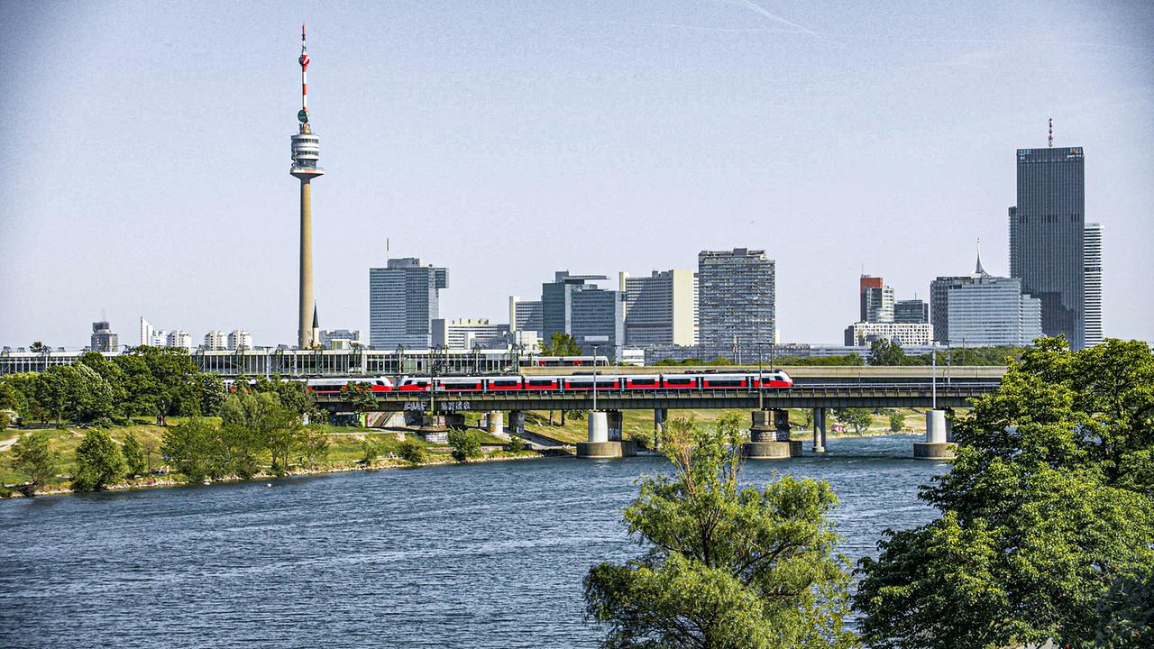 Die Wiener Skyline mit Donau, Donaubrücke, einem roten Zug und dem Donauturm im Hintergrund