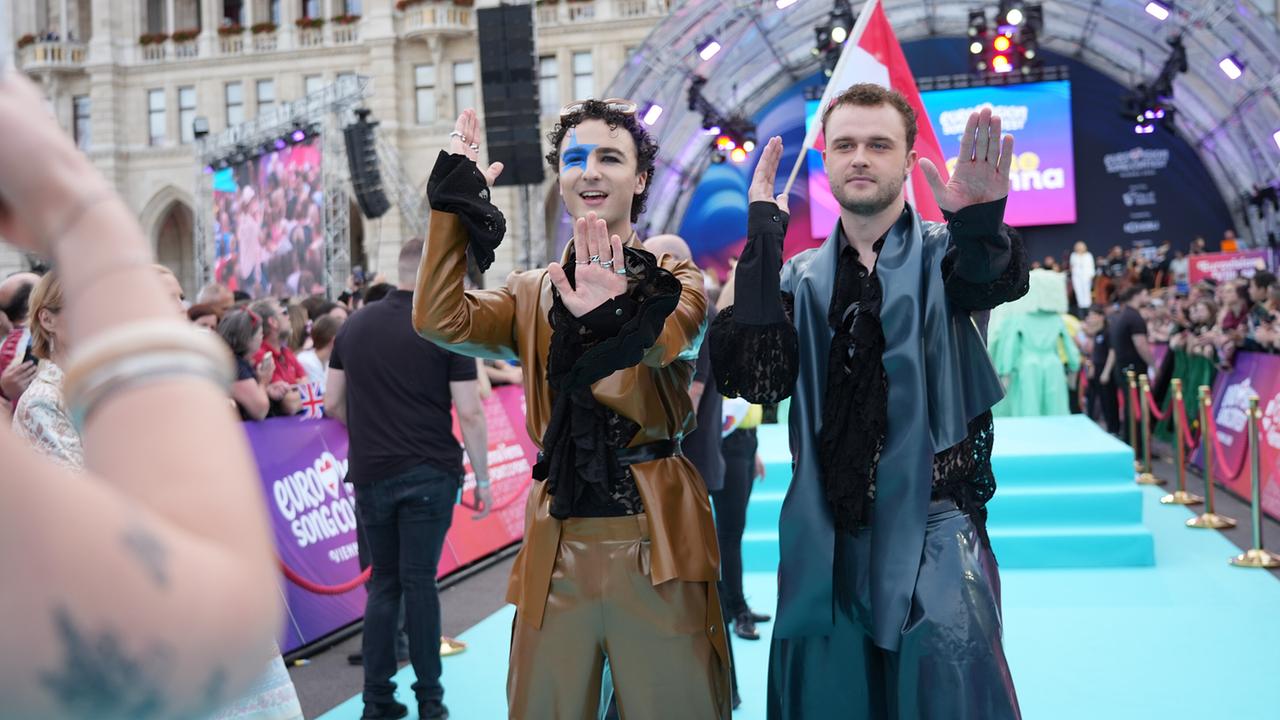 © ORF/Roman Zach-Kiesling COSMÓ and Sandro Humitsch perform "TANZSCHEIN" movements on the turquoise carpet at the grand ESC Opening in front of a large stage with an audience.