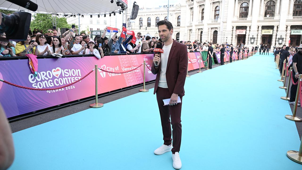© ORF/Hans Leitner Philipp Maschl stands on a turquoise carpet holding a microphone at a large public event, while a cheering crowd lines the barriers in front of an ornate historic building during the ESC Opening
