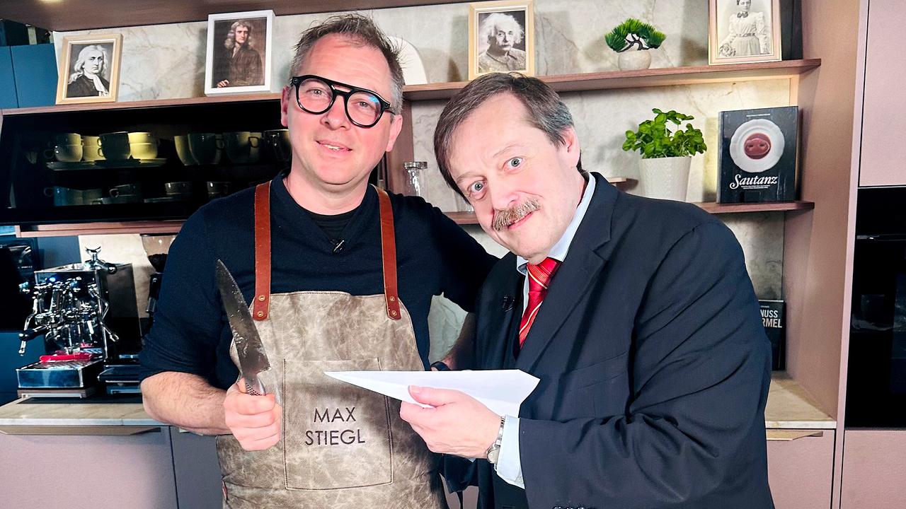 Max Stiegl mit einem Messer in der Hand und Werner Gruber mit einem Papierflieger in der Hand stehen nebeneinander in einer modernen Studioküche.