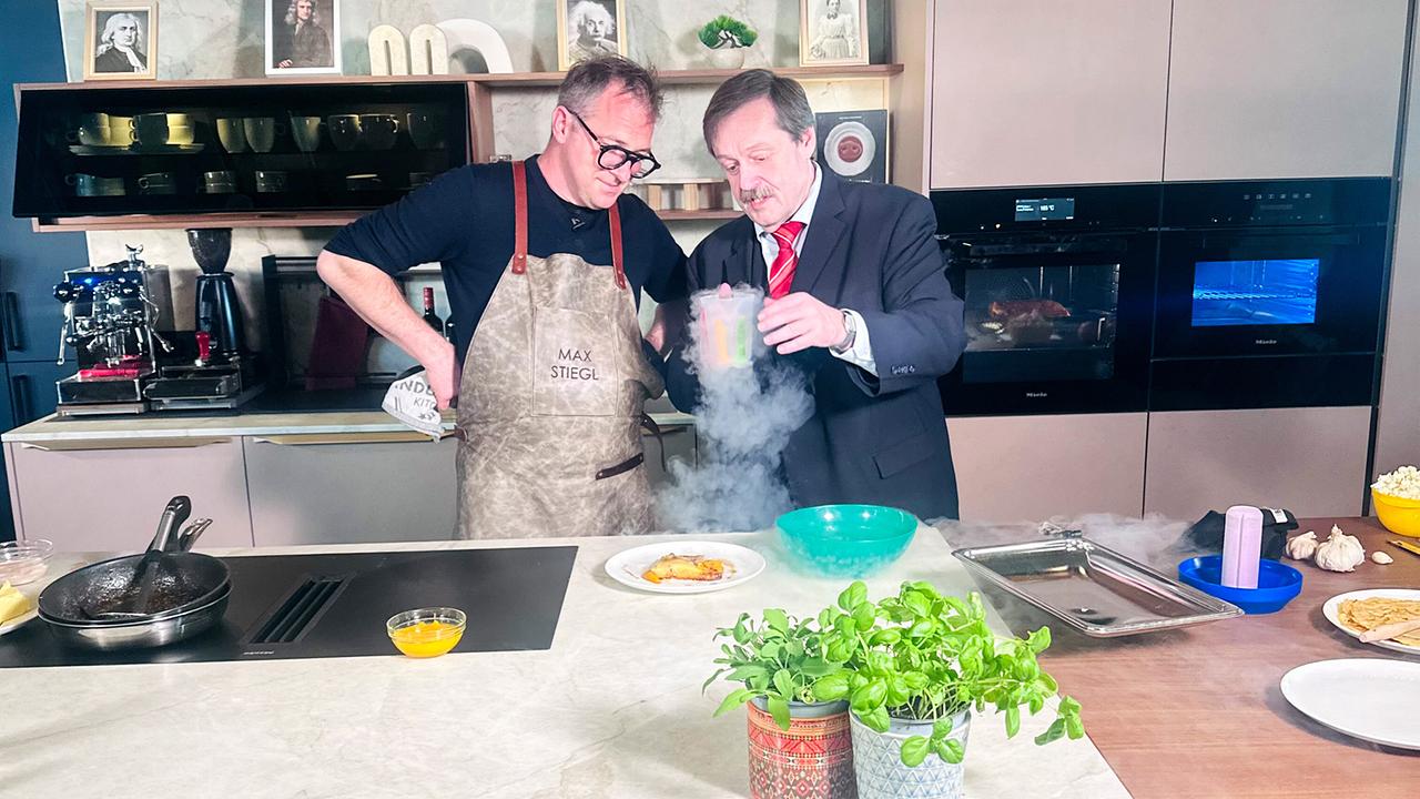 Max Stiegl und Werner Gruber stehen in einer modernen Studioküche, während Werner Gruber ein Gefäß hält, aus dem dichter Dampf aufsteigt.