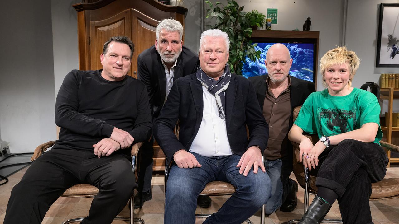Gruppenfoto mit Andi Herzog, Dirk Stermann, Toni Polster, Christoph Grissemann und Anna Mabo im "Willkommen Österreich"-Studio umgeben von Möbeln, Pflanzen und einem Aquarium im Hintergrund.