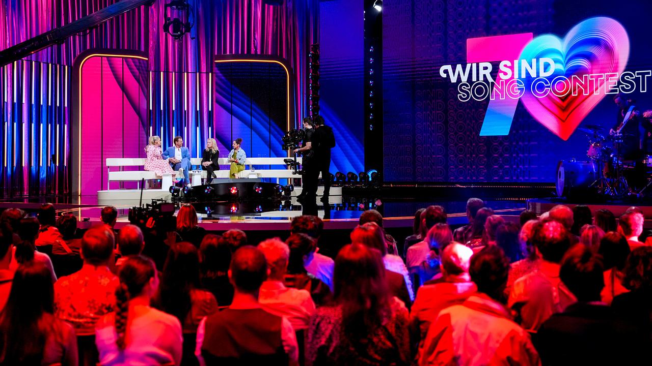 © ORF/Roman Zach-Kiesling Barbara Schöneberger, Mark Seibert, Nicole und Caroline Athanasiadis sitzen auf einer Coach auf der Bühne von "Wir sind Song Contest", mit Publikum im Vordergrund und Kameramännern auf der bunten Bühne.