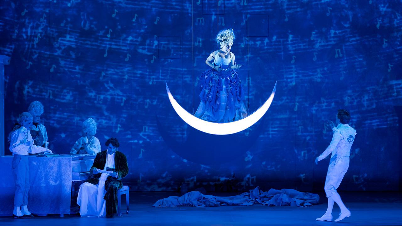 © ORF/Internationale Stiftung Mozarteum/Werner Kmetitsch In blauem Bühnenlicht schwebt die Königin der Nacht)in einem opulenten Kleid auf einer leuchtenden Mondsichel, während Tamino und weitere Figuren darunter wie in einer Traumsequenz agieren.