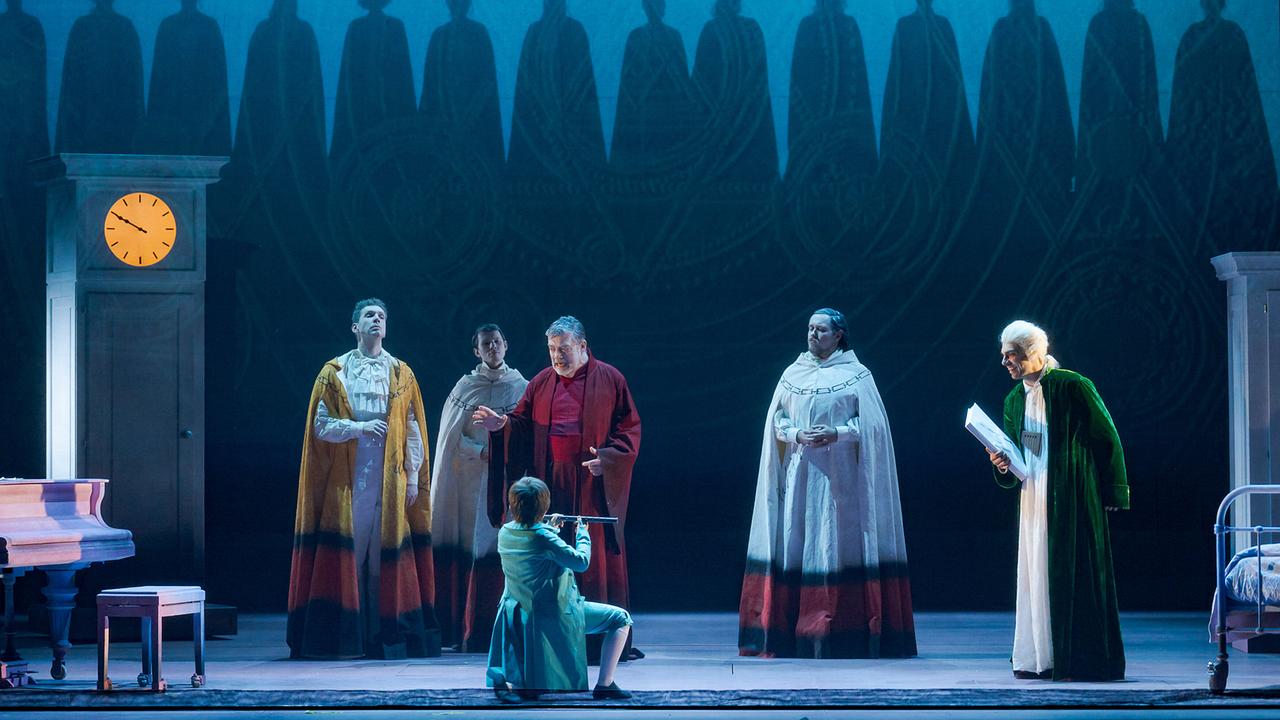© ORF/Internationale Stiftung Mozarteum/Werner Kmetitsch Vor dem Bühnenbild einer Reihe schattenhafter Gestalten stehen mehrere Figuren in historischen Gewändern unter ihnen Sarastro und Mozart, ein Kind kniet mit einer Flöte davor, links ist eine Uhr und ein Klavier zu sehen.