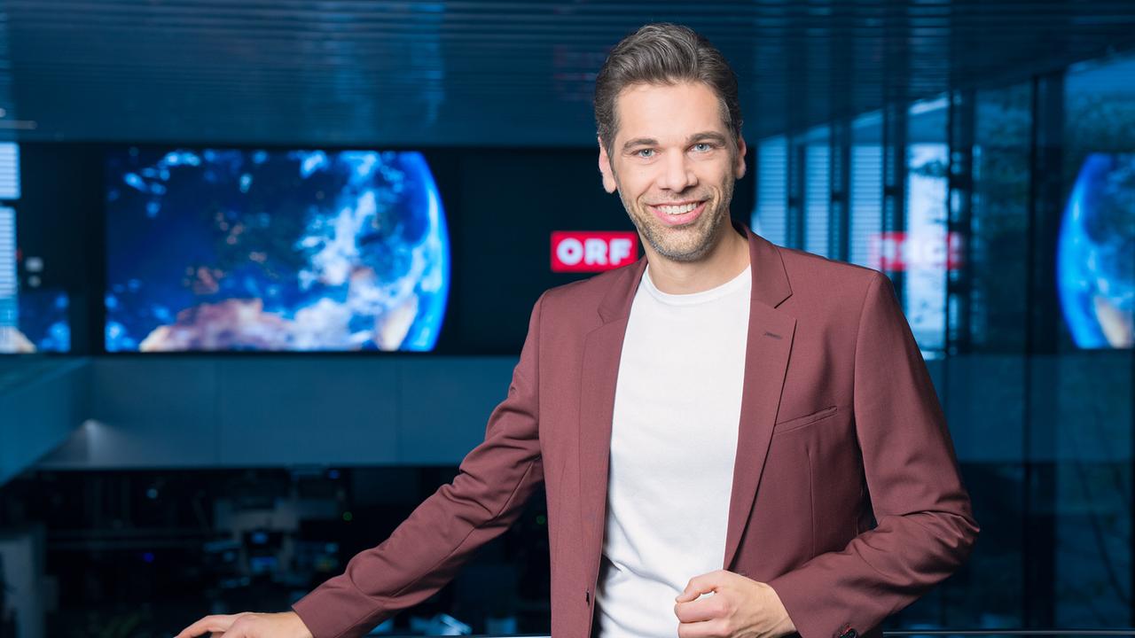 Philipp Maschl in einem bordeauxfarbenen Anzug und weißem Shirt steht im ORF-Newsroom vor großen Bildschirmen mit einer Weltansicht.