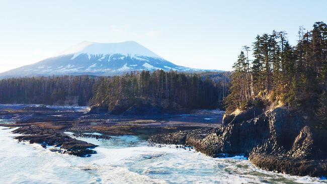 Meer und Felsen mit Nadelbäumen, im Hintergrund ein schneebedeckter Berg, könnte ein Vulkan sein.