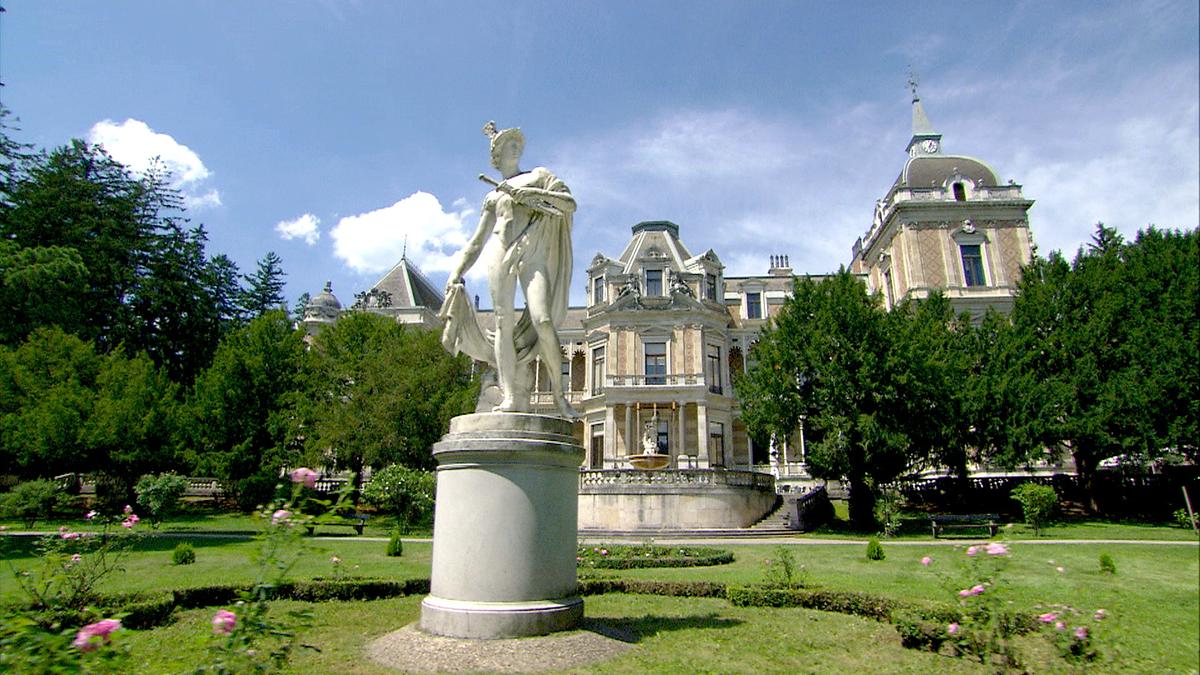 © ORF/Rilk Film Die Statue des griechischen Gottes Hermes aus weißem Marmor im Garten vor der Hermesvilla. Das Gebäude ist von Bäumen eingerahmt, im Vordergrund bei der Statue Wiese mit einigen rosa blühenden Rosensträuchern.