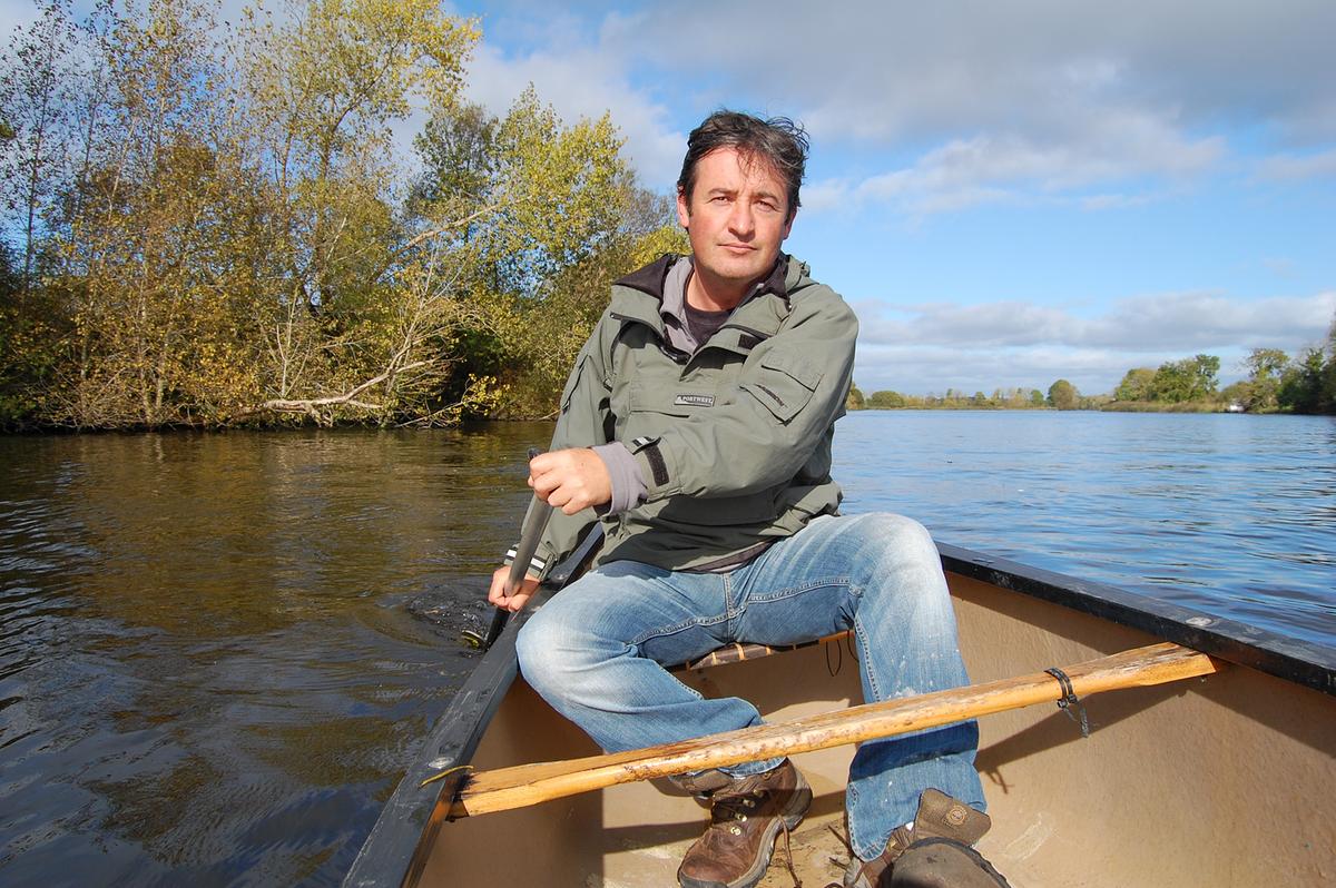 Der Moderator Colin Stafford Johnson rudert in einem Boot am Fluss Shannon. Im Hintergrund Bäume und Sträucher, bewolkter Himmel.