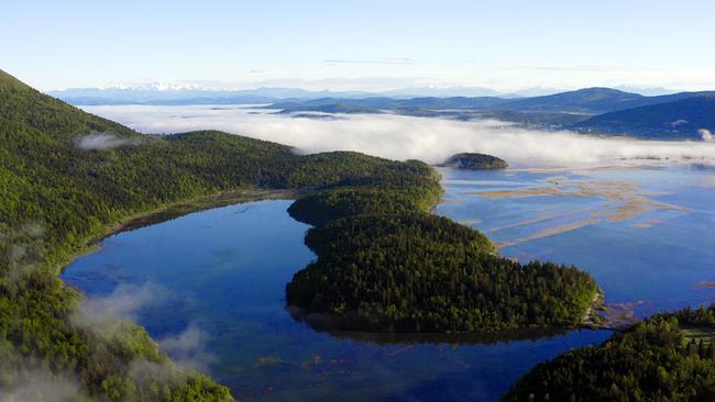 Ein blau schimmernder See mit perlenstrangförmig angeordneten Inseln, Ufer und Inseln sind grün bewachsen. Im Hintergrund Nebel und Berge.