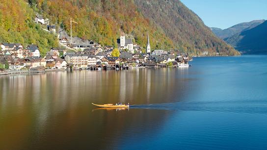 Ein Boot fährt im Hallstättersee, links im Bild schmiegt sich Hallstatt an den Berghang. Der Wald an den steilen Uferhängen sieht herbstlich aus.
