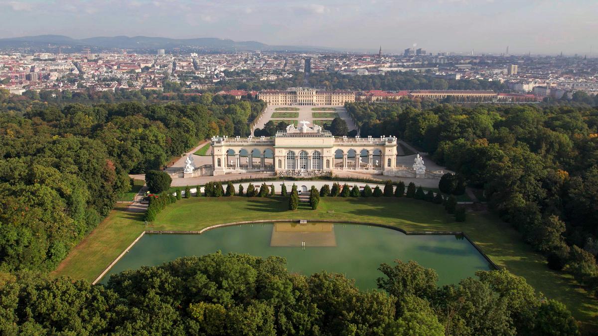 Die Gloriette mit Teich im Vordergrund, in der Blickachse dahinter erst das barocke Schloss Schönbrunn und dann die Stadt Wien.