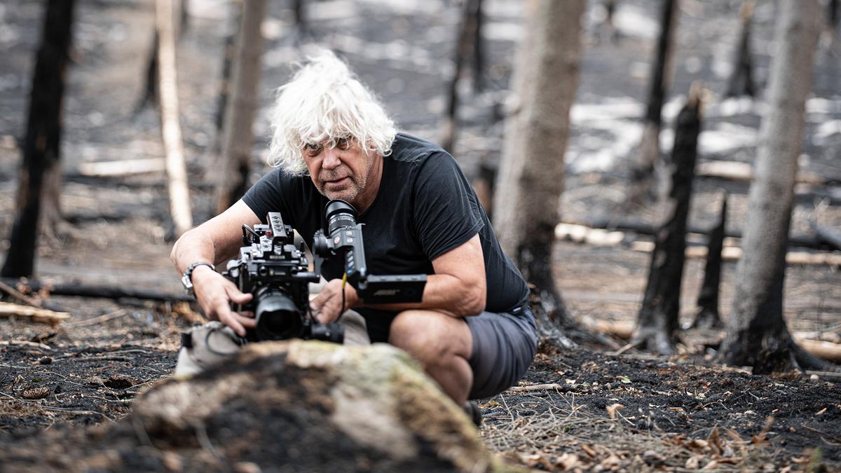 Ein älterer Mann mit weißen, kinnlangen Haaren und Dreitagebart kniet mit der Kamera am Boden. Er trägt ein schwarzes T-Shirt und kurze schwarze Hosen. Der Waldboden ist schwarz verbrannt, auch die Baumstämme weisen Brandspuren auf.