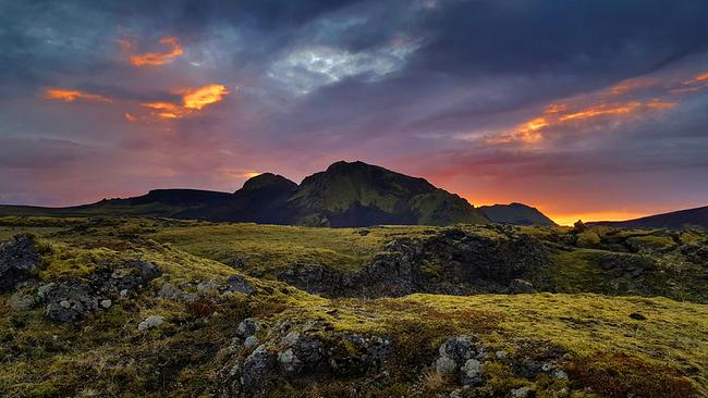 Schroffe Felsen mit kurzem grünen Bewuchs, im Hintergrund Berge. Die Wolken leuchten intensiv in orange, violett und blau.
