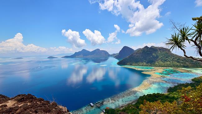 Eine Gruppe von grünen Hügeln mit steilen Hängen, die aus dem türkisblauen Wasser ragen. Wenige Wolken am strahlend blauen Himmel, sie spiegeln sich im glatten Meer. Den Vordergrund des Fotos bilden schroffe Felsen.