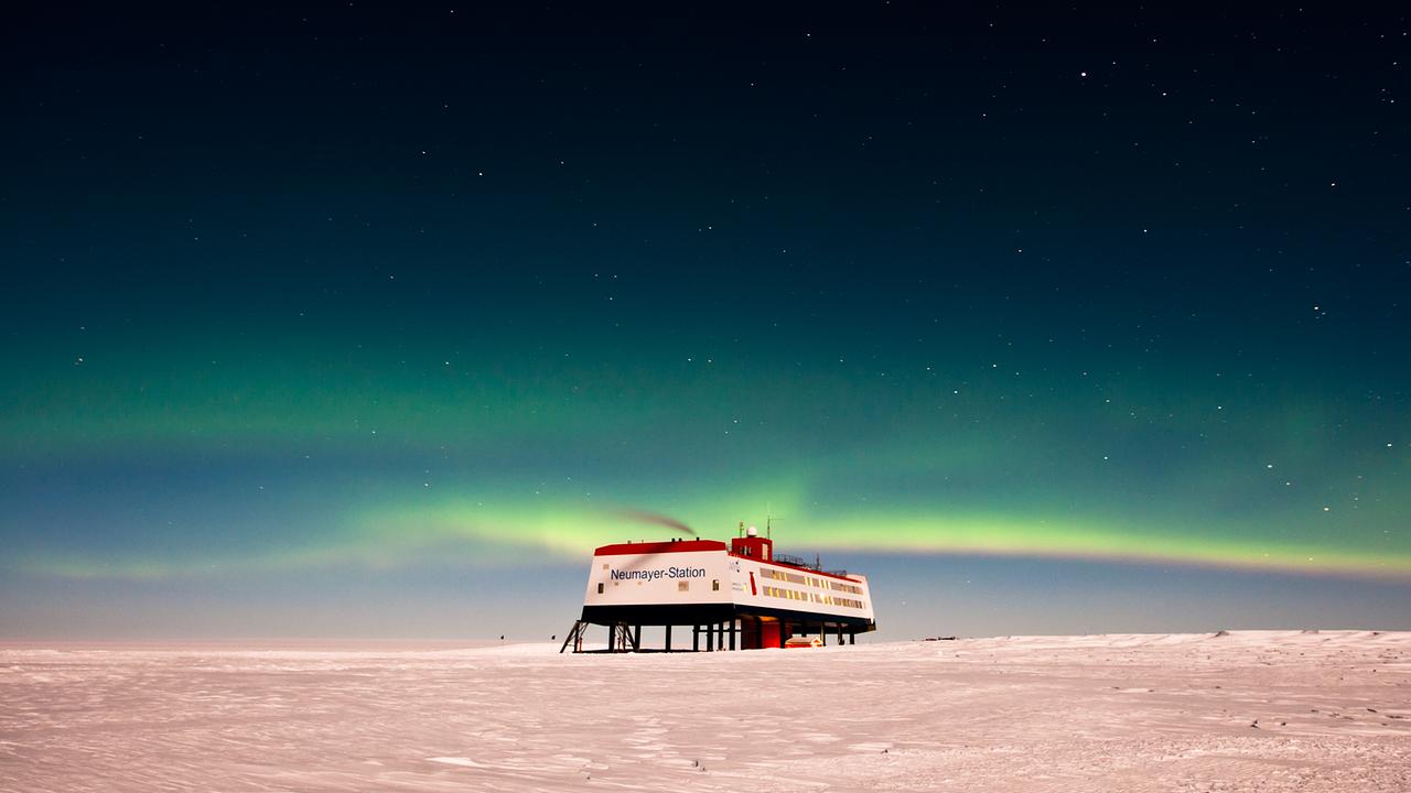 Die deutsche Antarktis-Forschungsstation Neumayer-Station III wirkt klein auf der schneebedeckten Ebene. Es ist Nacht, limettengrüne Polarlichter sind am Himmel zu erkennen.