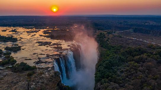 Sonnenuntergangsstimmung an den Victoriafällen, der Sambesi ist links im Bild. In der Mitte stürzt er über die Kante und bildet Sprühnebel. Rechts befindet sich dichter Wald.