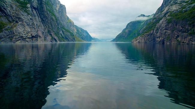 Ein Fjord in Norwegen, vom Wasser aus fotografiert. Die steilen, gegenüber liegenden Felswände mit wenig grünem Bewuchs bilden durch die niedrige, mittige Kameraposition ein V. Dichte Wolken am Himmel, das Wasser zwischen den Felswänden ist ruhig. Himmel und Gestein spiegeln sich an der Wasseroberfläche.