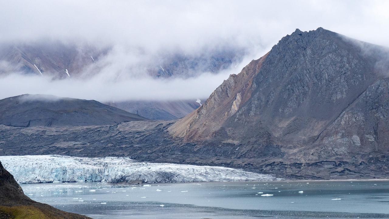 Ein Meeresarm mit einem Gletscher, im Hintergrund befindet sich ein schroffer, kegelförmiger Berg. Das übrige Gebirge wird von Nebel eingehüllt. Die kurze Vegetation ist bräunlich-grün, das Wasser stahlblau.