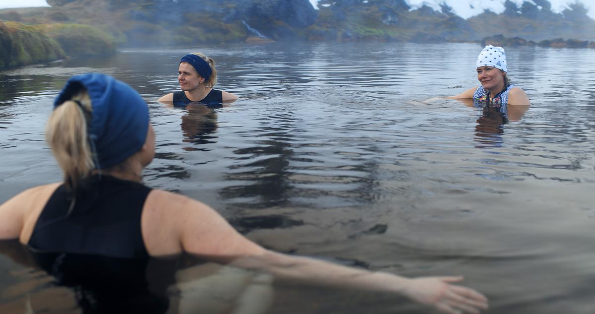 Drei Frauen mit Kopfbedeckungen sitzen im flachen Wasser einer heißen Quelle. Der Bewuchs am Rand ist bräunlich-grün, Schneereste sind vorhanden.