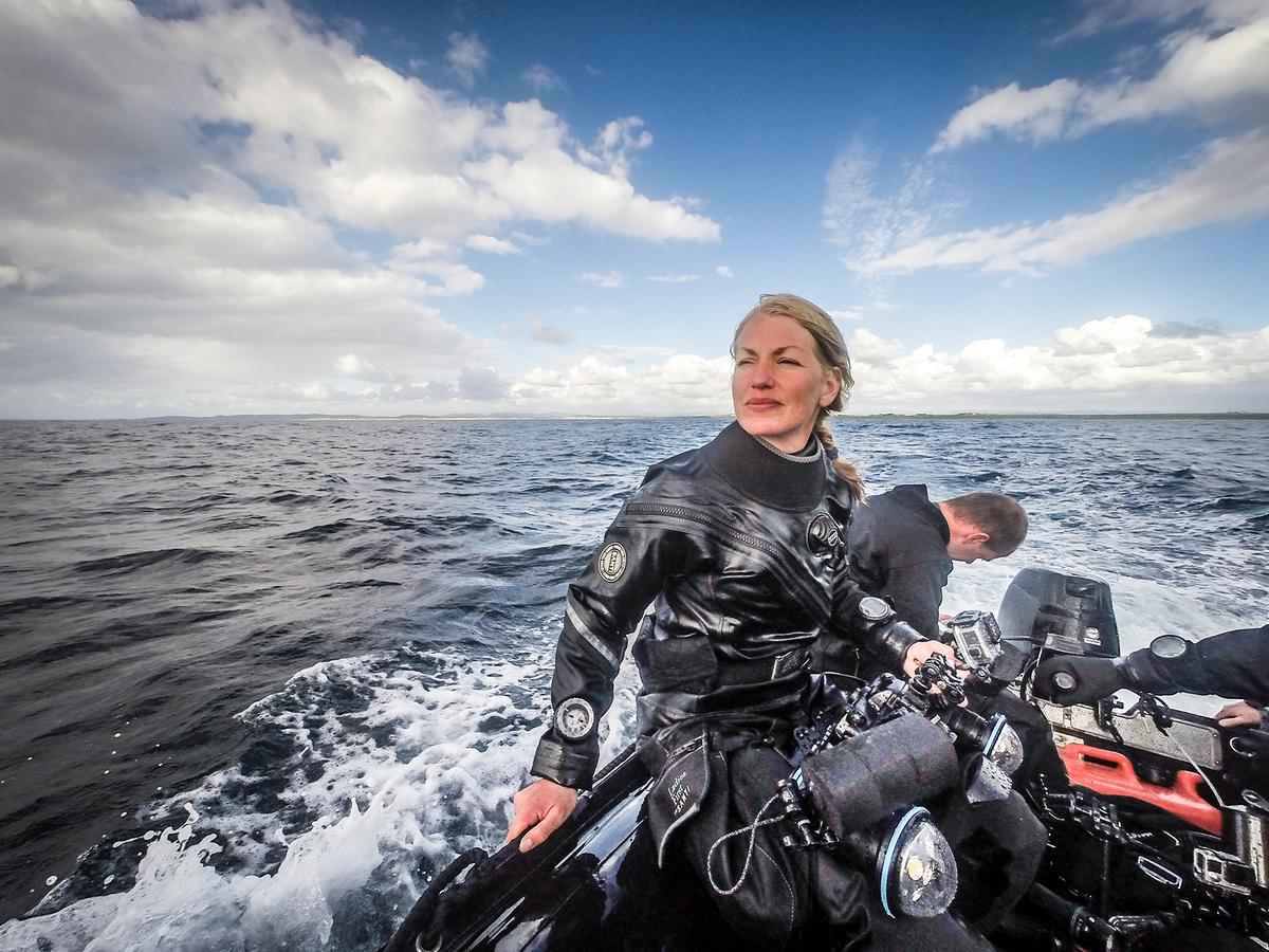 Die Regisseurin im Taucheranzug auf einem Motorboot, das am offenen Meer fährt. Sie hält ihre Kamera am Schoß, im Hintergrund ein Kollege. 