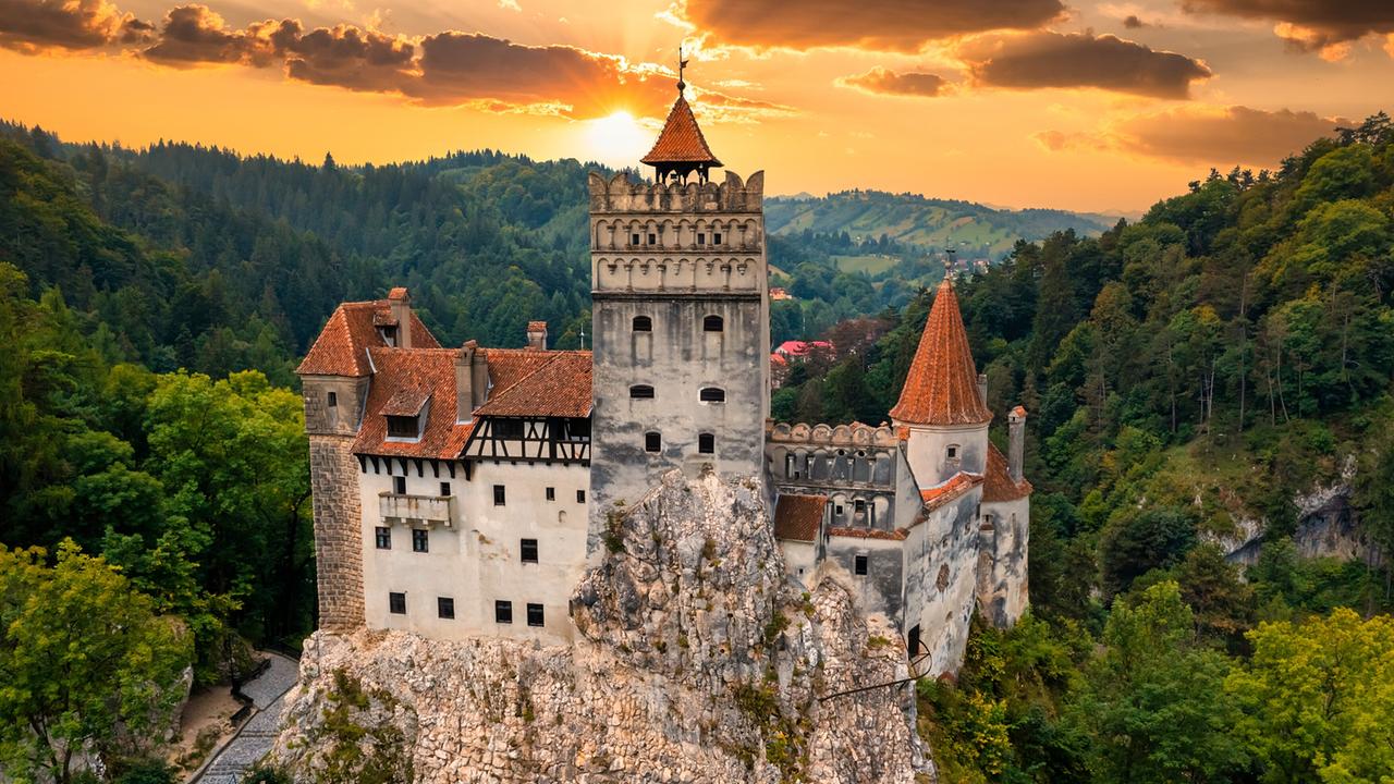 © ORF/Crossing the Line/fogcatcher/Shutterstock Ein Schloss auf einem Felsen, umgeben von grünen Wäldern. Der Himmel mit tiefstehender Sonne ist orange.