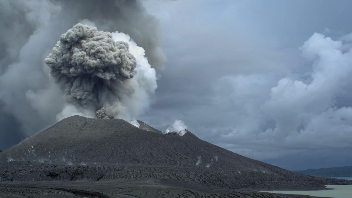 Der schwarze Krater eines Vulkans, eine große Aschewolke steigt daraus auf.