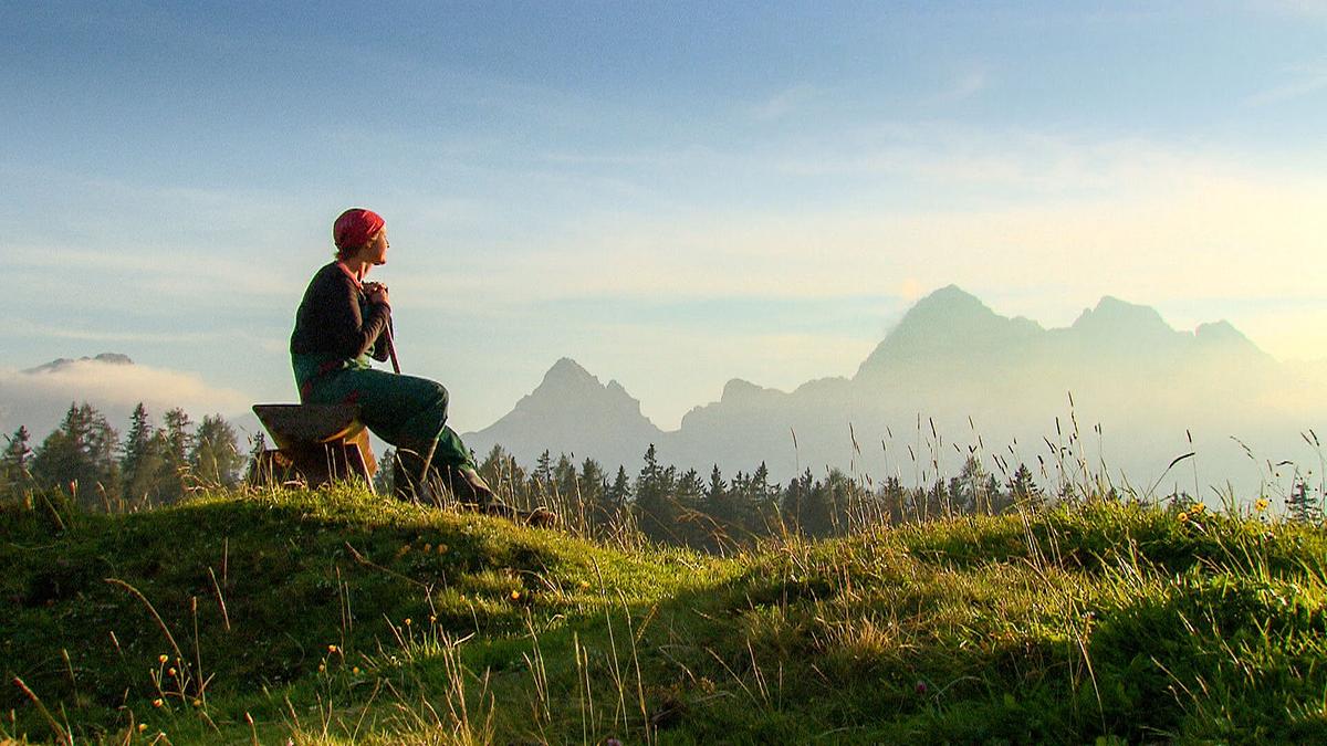 Eine Sennerin mit rotem Kopftuch sitzt auf einer Holzbank, sie stützt sich auf einen Wanderstock und schaut in die Ferne. Ein Gebirgskamm bildet den Hintergrund. Die Sonne steht tief und wirft lange Schatten.