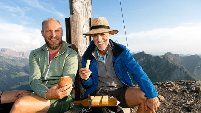 Hermann Maier hält Gebäck, Toni Innauer ein Stück Bergkäse. Sie sitzen am Fuß des Gipfelkreuzes der Kanisfluh, das Schneidbrett mit dem Käse ruht auf einem Rucksack. Im Hintergrund steile Berge und blauer Himmel.