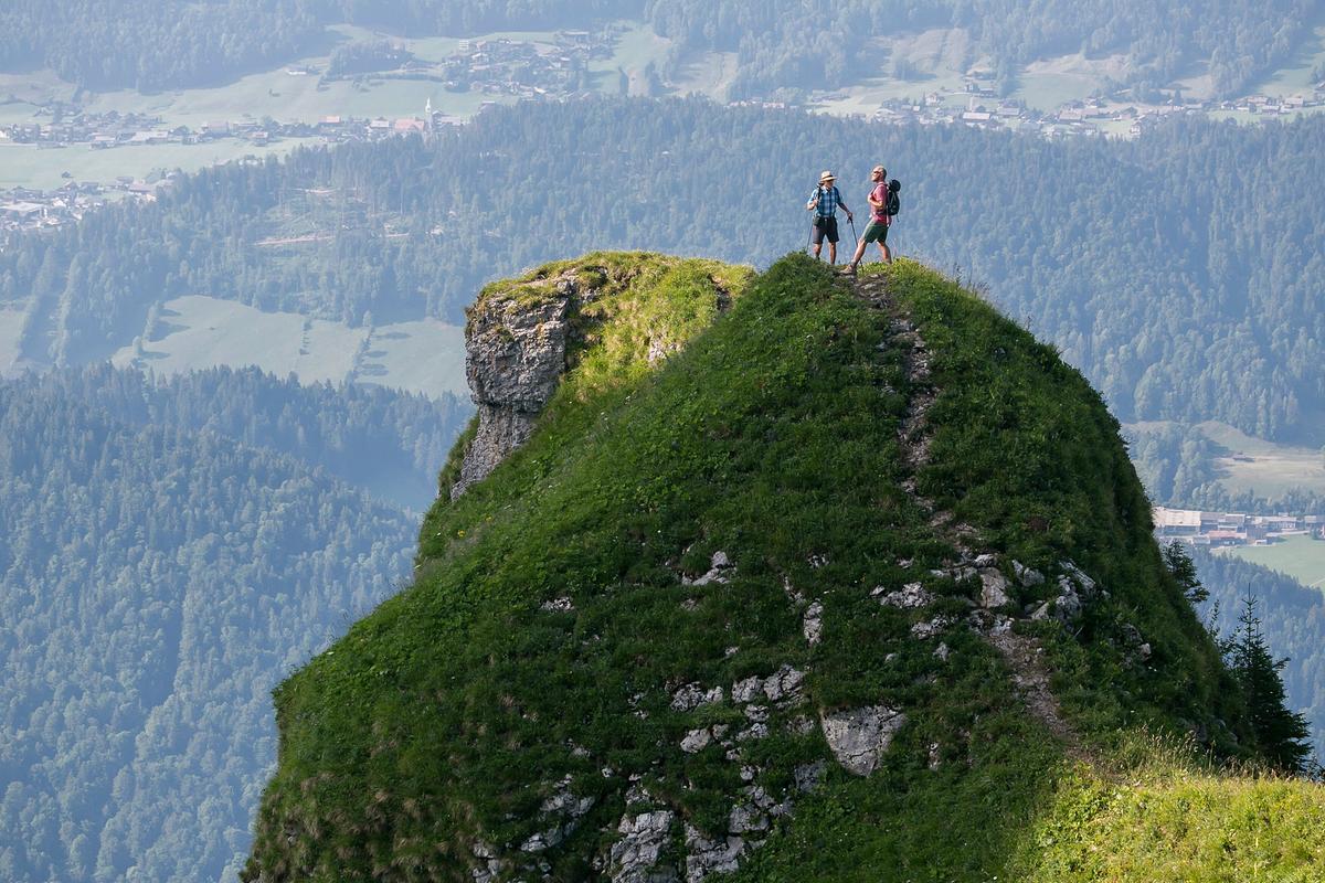 Hermann Maier und Toni Innauer stehen auf einem steilen, grün bewachsenen Felsen. Hinter ihnen liegt das Tal, dadurch wird die große Höhe der Kanisfluh deutlich.